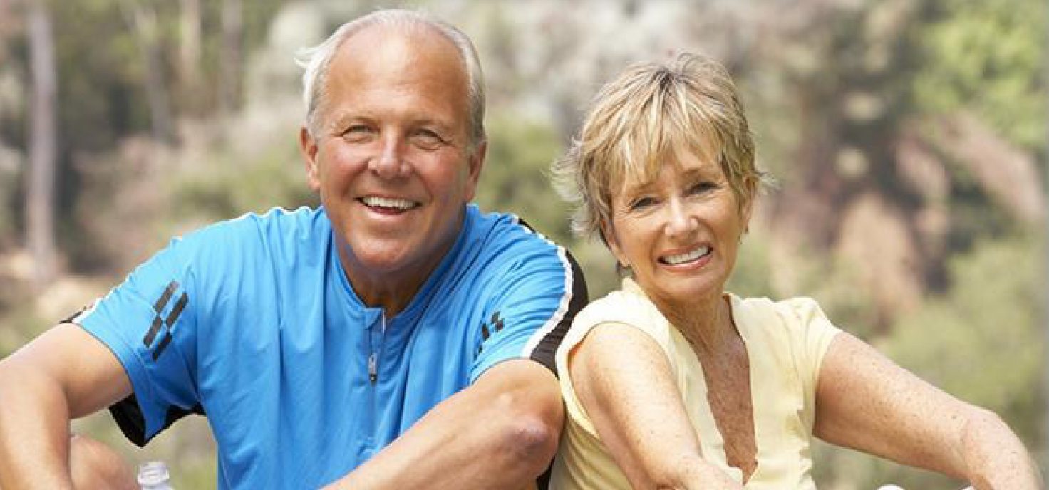 A smiling man and woman sitting in a park wearing workout clothing.